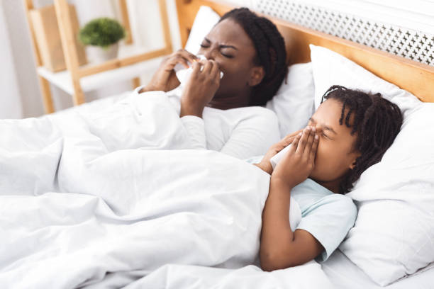 5 Respiratory Disease. Black mom and daughter sneezing, blowing noses on paper napkins in bed